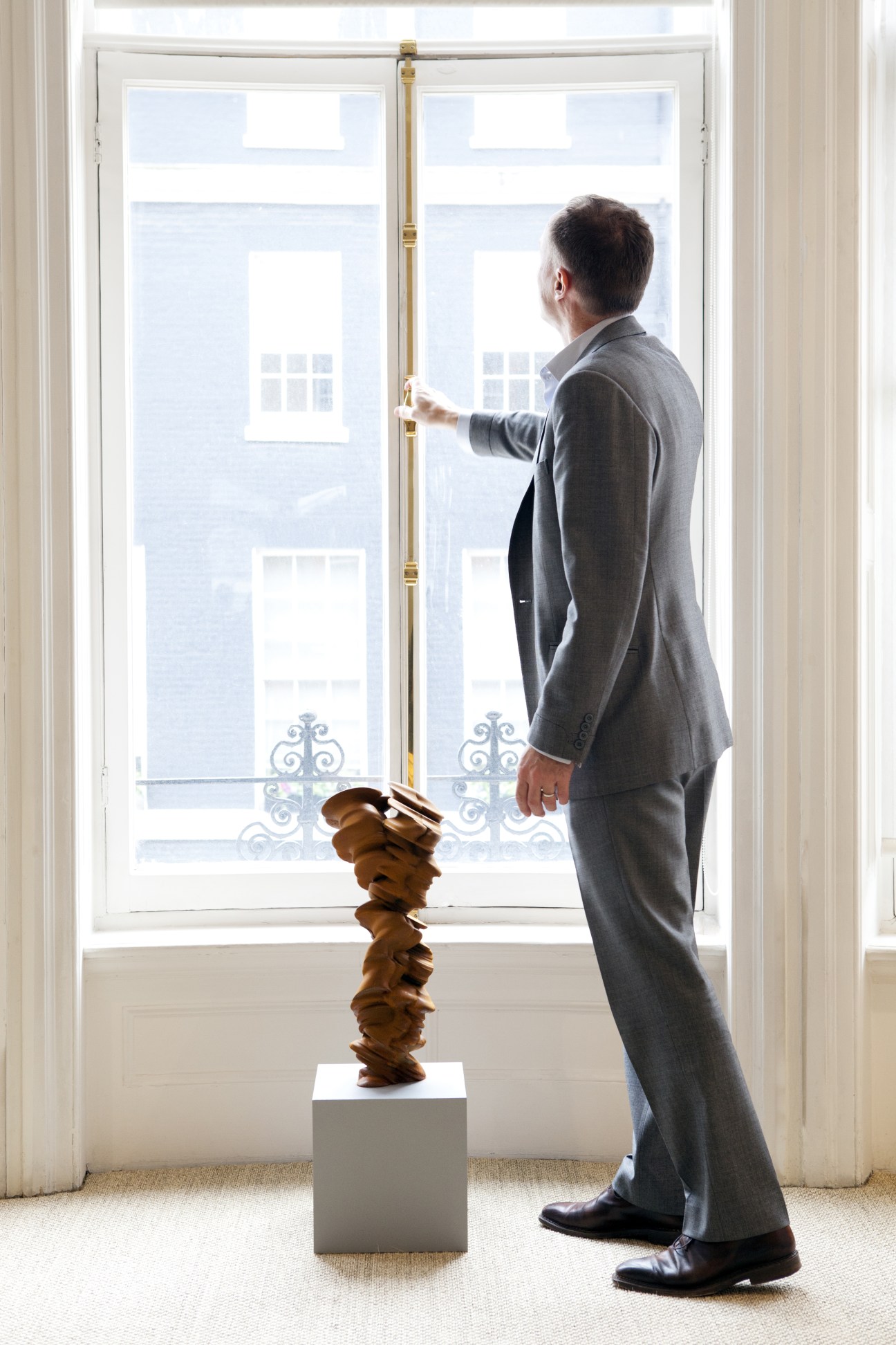 Gallery owner Harry Blain stands beside a wooden sculptural piece by Tony Cragg, gazing out tall windows.