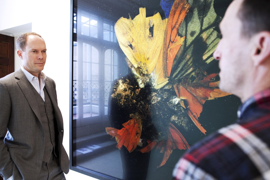 Gallery owner Harry Blain standing beside Mat Collishaw's 'Insecticide 3' artwork featuring colorful butterflies.