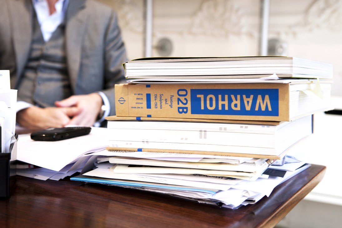 Stack of art sale catalogues and invitations on a desk with a man in business attire seated behind.
