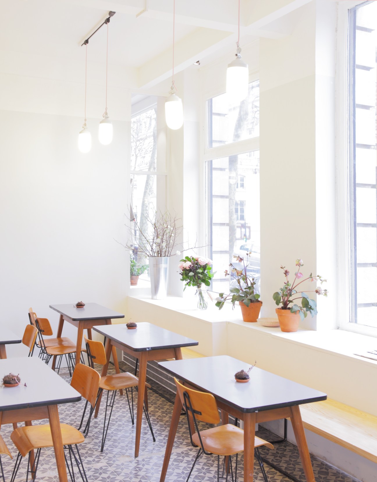 Bright café interior with mid-century wooden tables, chairs, hanging pendant lights, and potted flowers on windowsill.