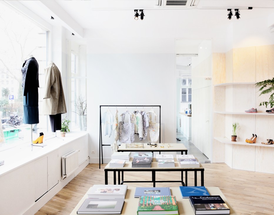 Minimalist fashion boutique interior with white walls, wooden floor, clothing displays, and magazines on tables.