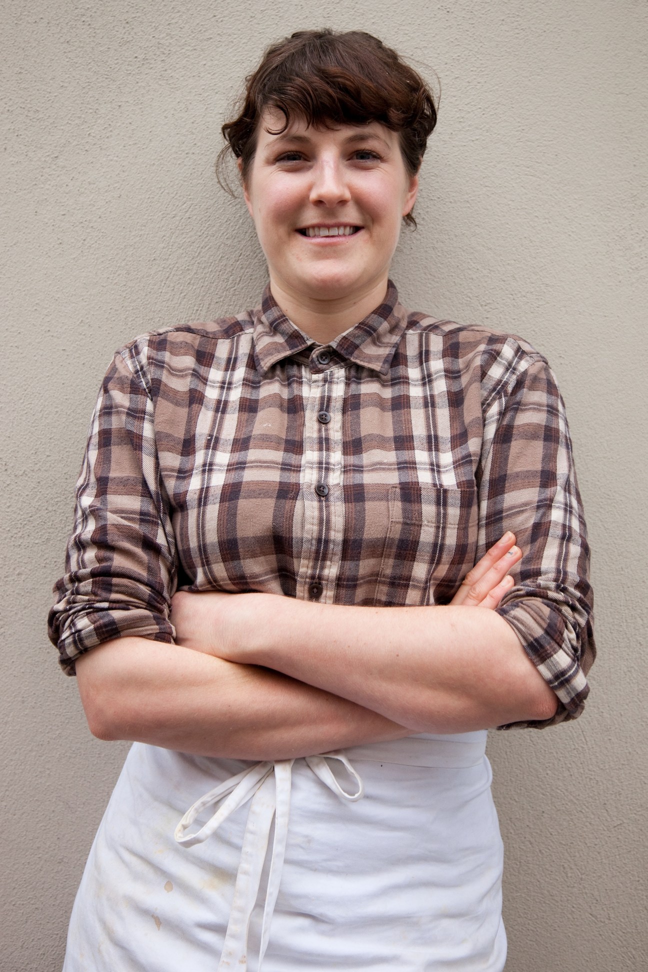 Woman wearing a plaid shirt and white apron smiles at the camera with arms crossed.