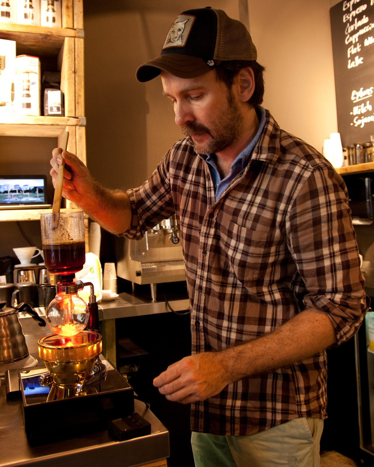 Man in plaid shirt and cap preparing coffee using a pour-over method at The Barn café in Berlin.