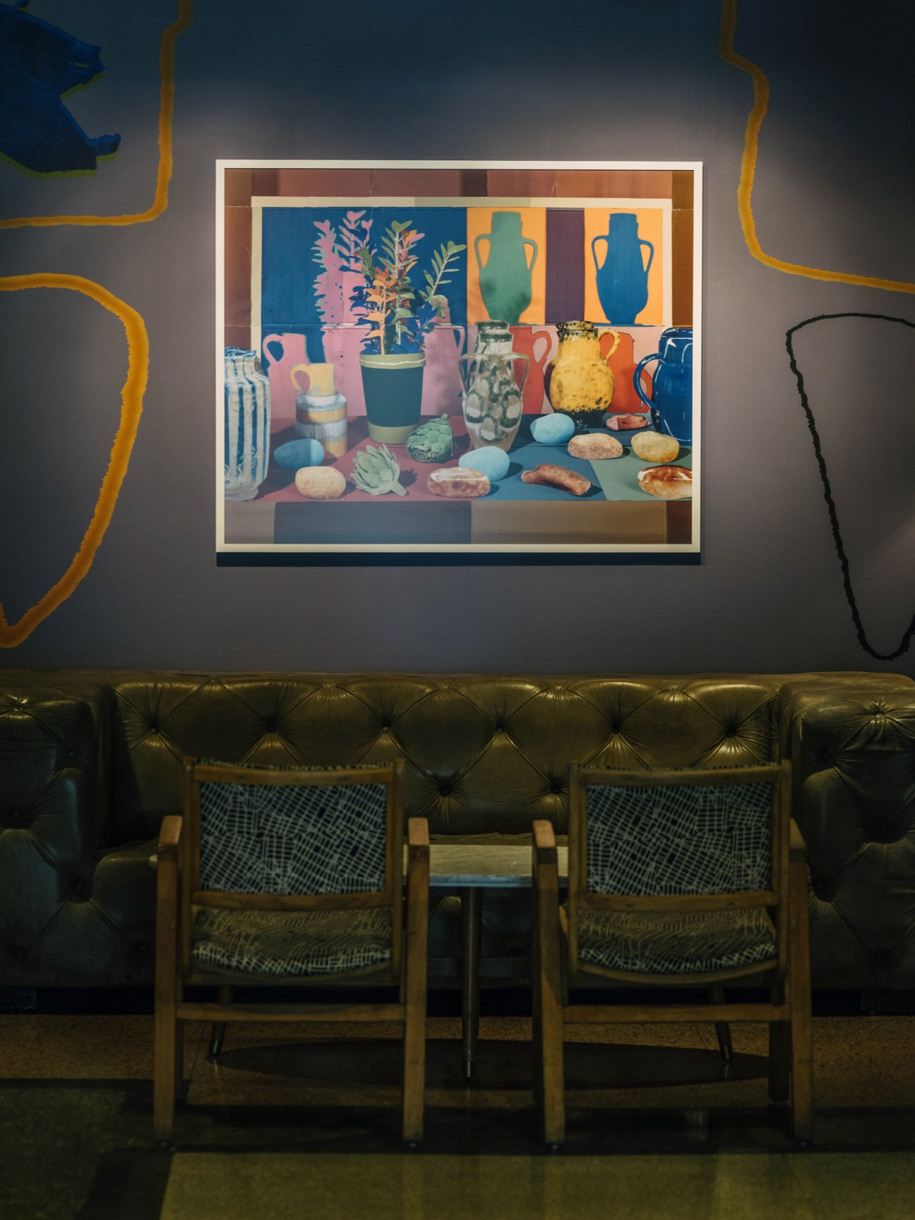 Colorful framed still-life artwork featuring vases, plants and objects displayed on a gallery wall above a black leather sofa.