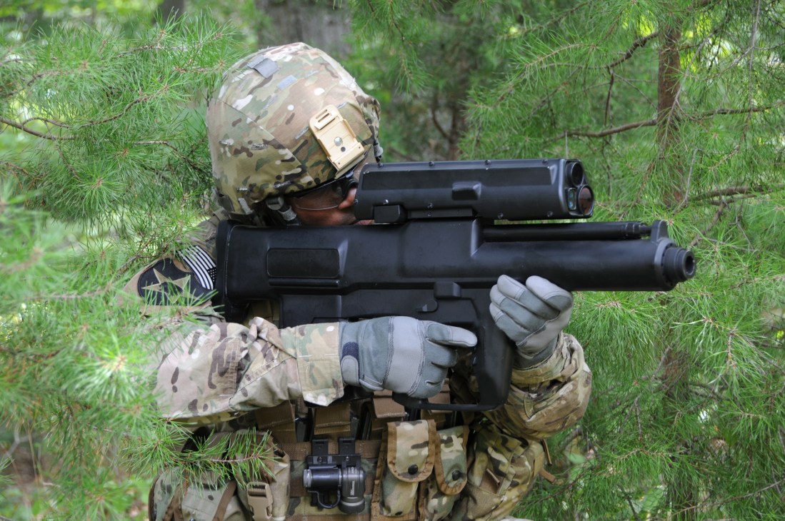 US soldier in camouflage uniform holding XM25 grenade launcher in forest setting.