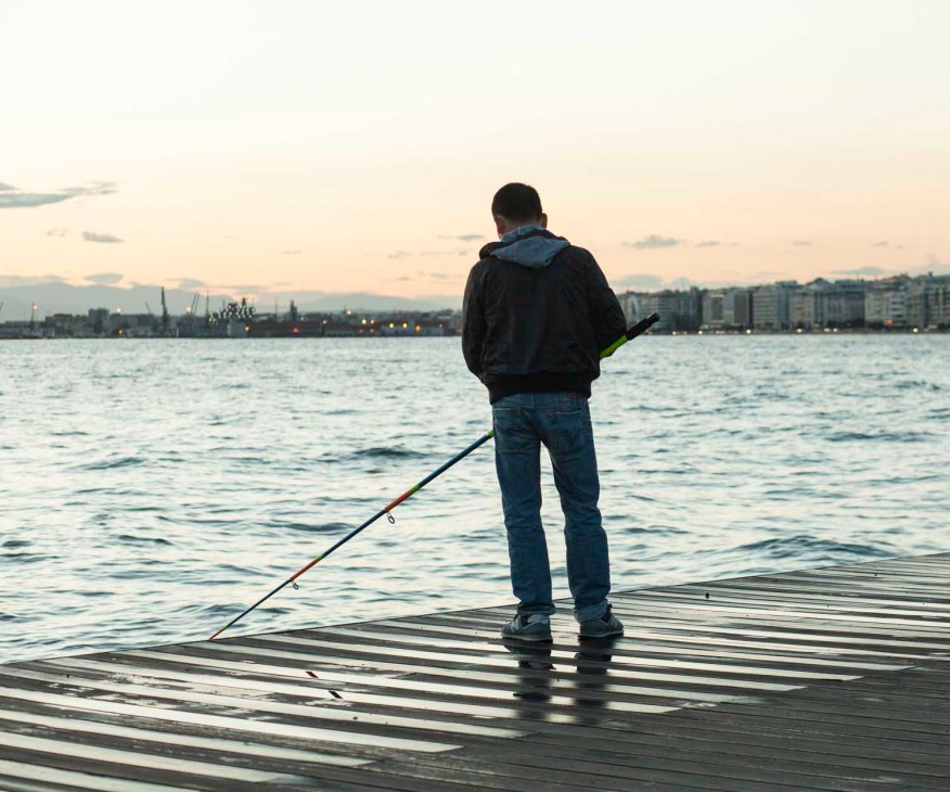Person fishing on a wooden pier at sunset overlooking Thessaloniki's waterfront and cityscape.