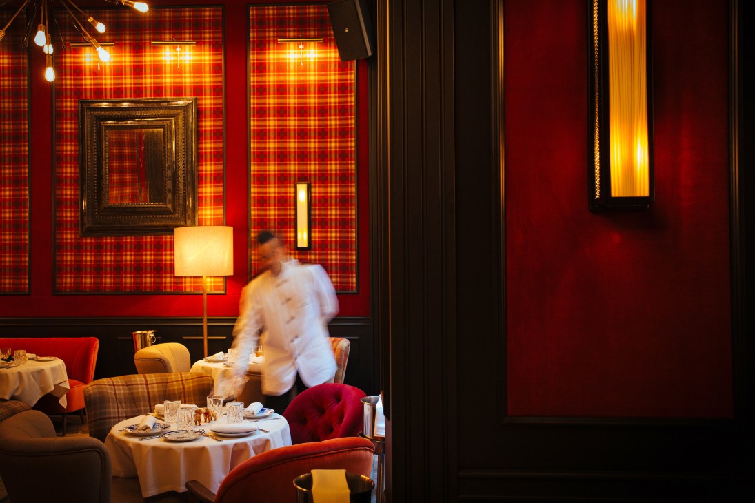 Server in white shirt moves through The Wittmore Hotel's dining room with red tartan wallpaper and warm lighting.