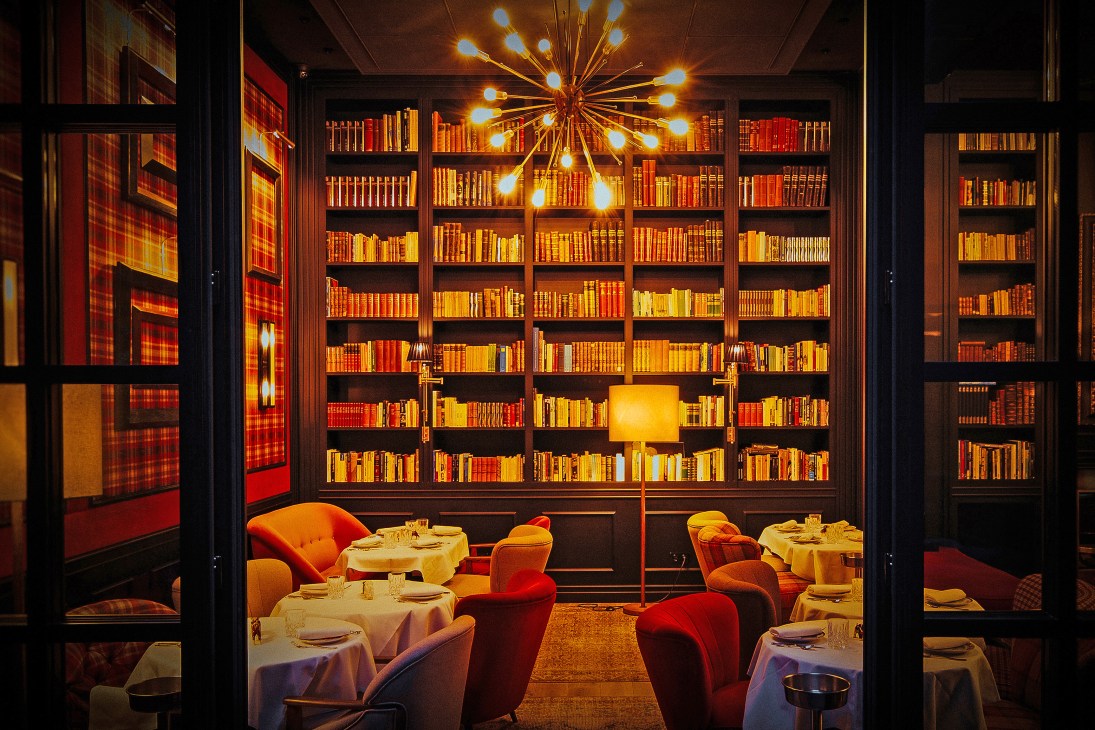 The Wittmore Hotel's dining room with floor-to-ceiling bookshelves, starburst chandelier, and red velvet chairs at candlelit tables.