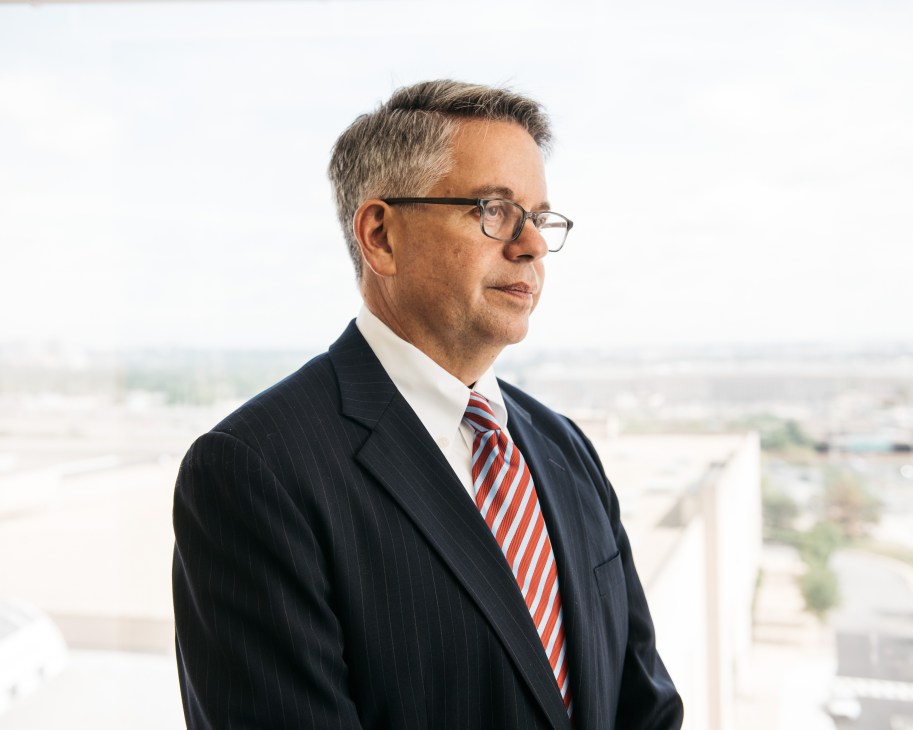 Man in business suit and striped tie stands before a window overlooking a cityscape.