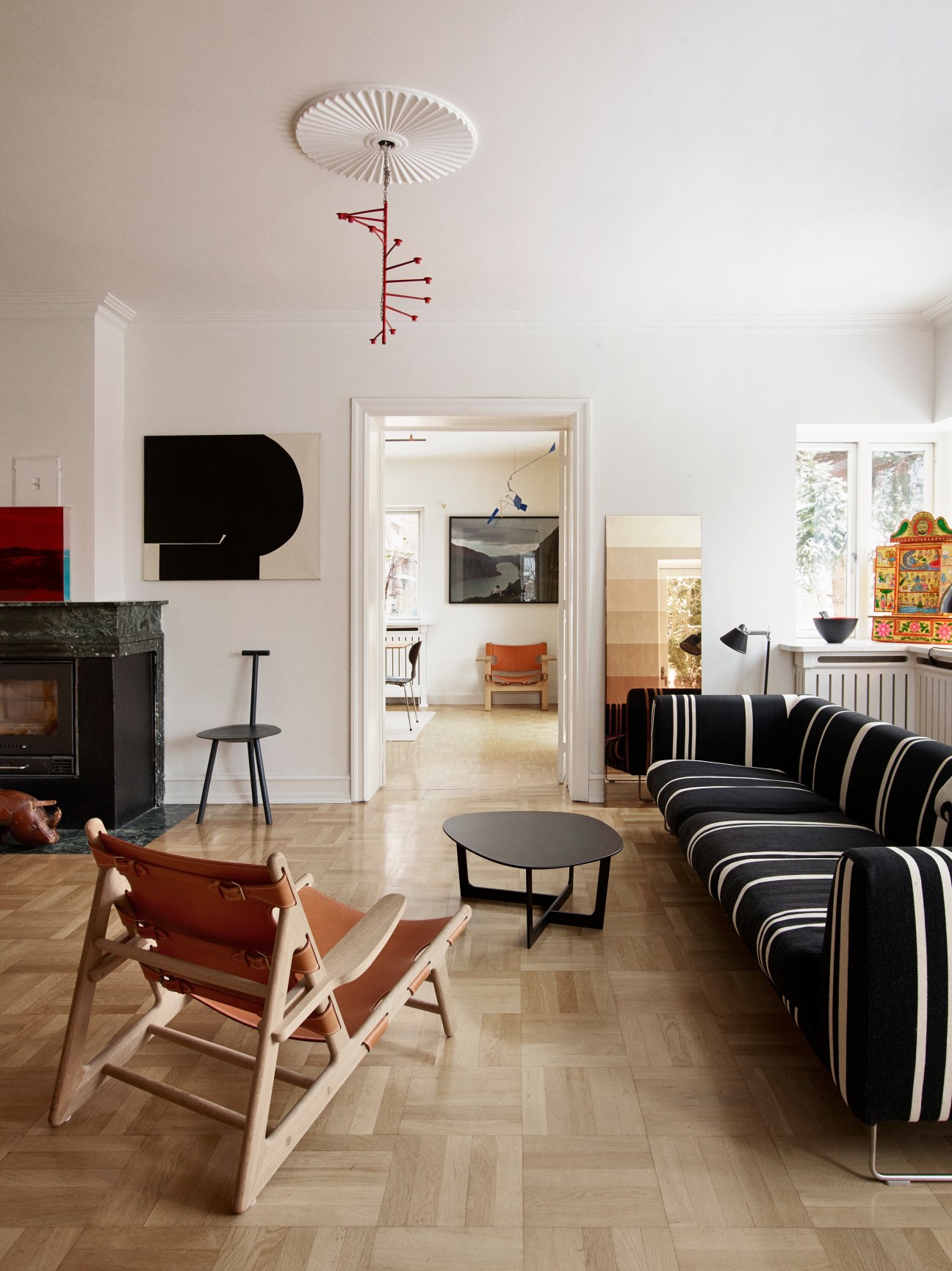 Modern living room with Børge Mogensen's Hunting Chair in cognac leather, striped black and white sofa, red mobile art installation, and minimalist Danish furniture.