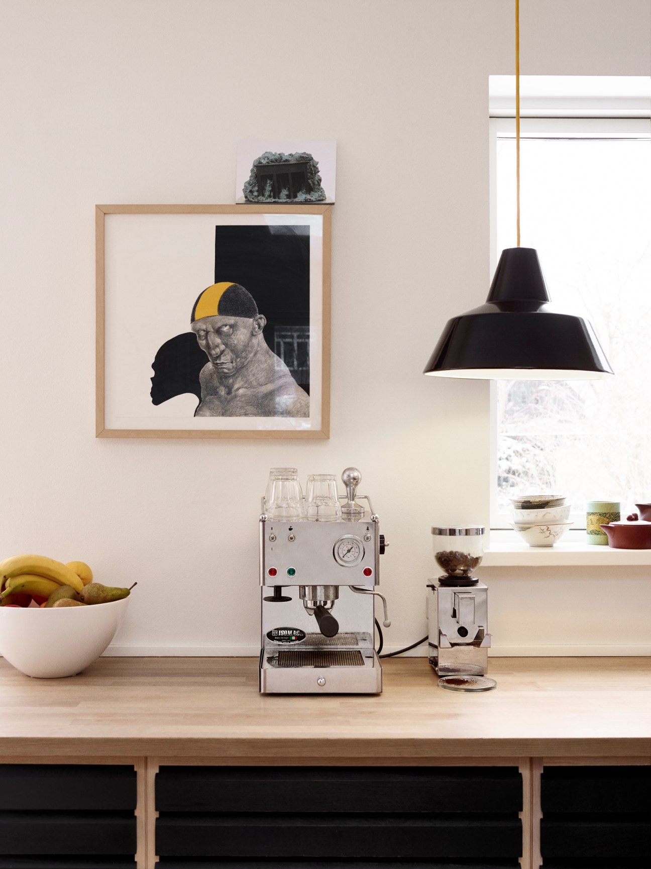 Kitchen counter displaying a Michael Kvium print, espresso machine, black pendant lamp, and bowl of bananas.
