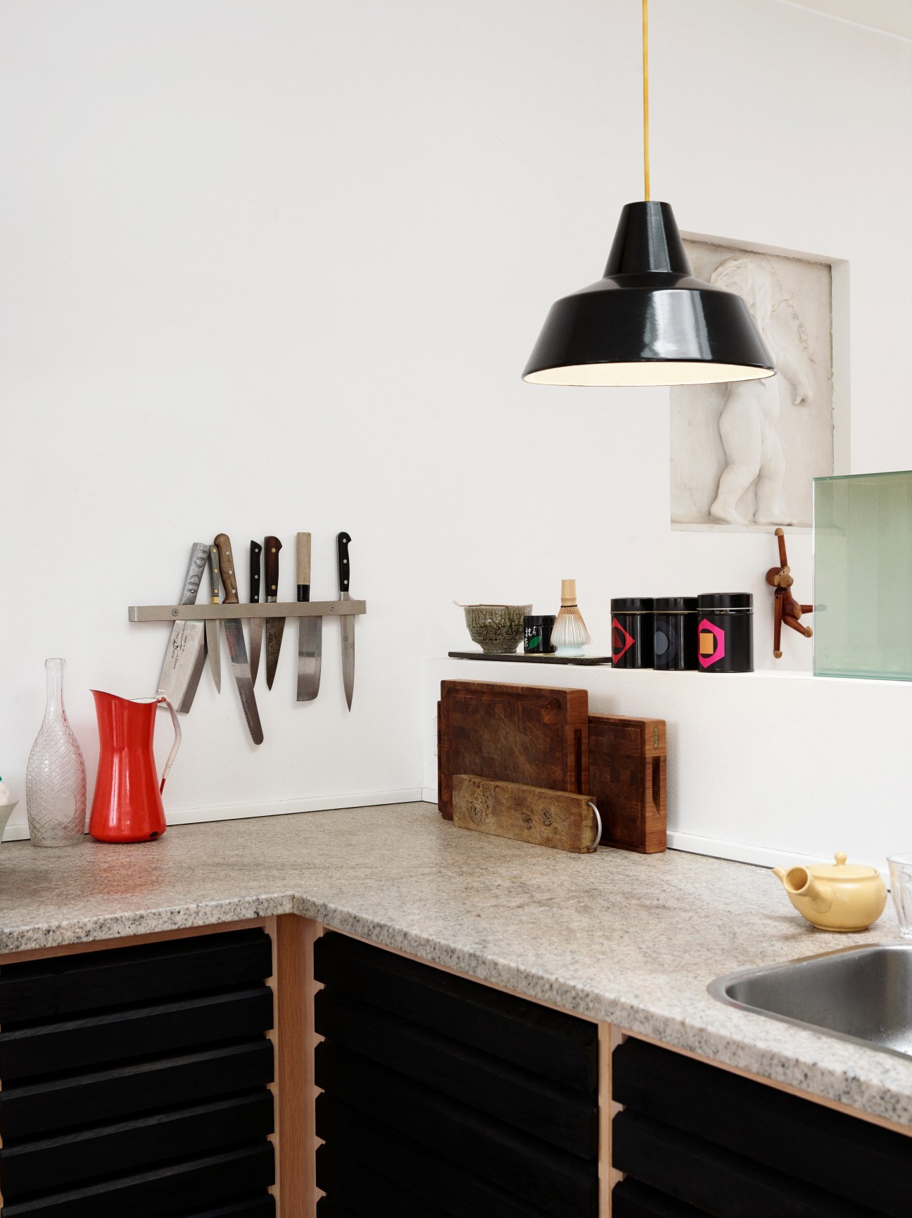 Kitchen counter with black pendant lamp, knife rack, red pitcher, and wooden design objects on white walls.