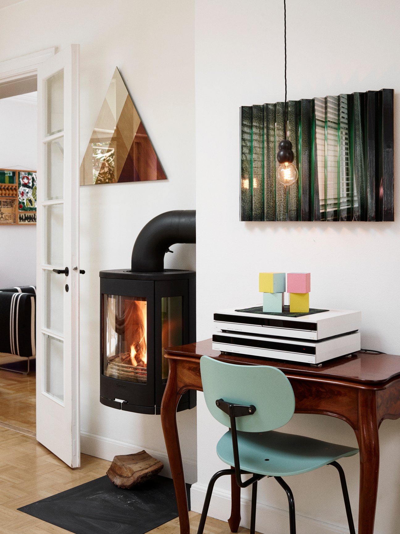Interior room with wooden desk, mint green Egon Eiermann chair, stacked books, colorful cubes, fireplace, and abstract artwork by Mette Kjær.