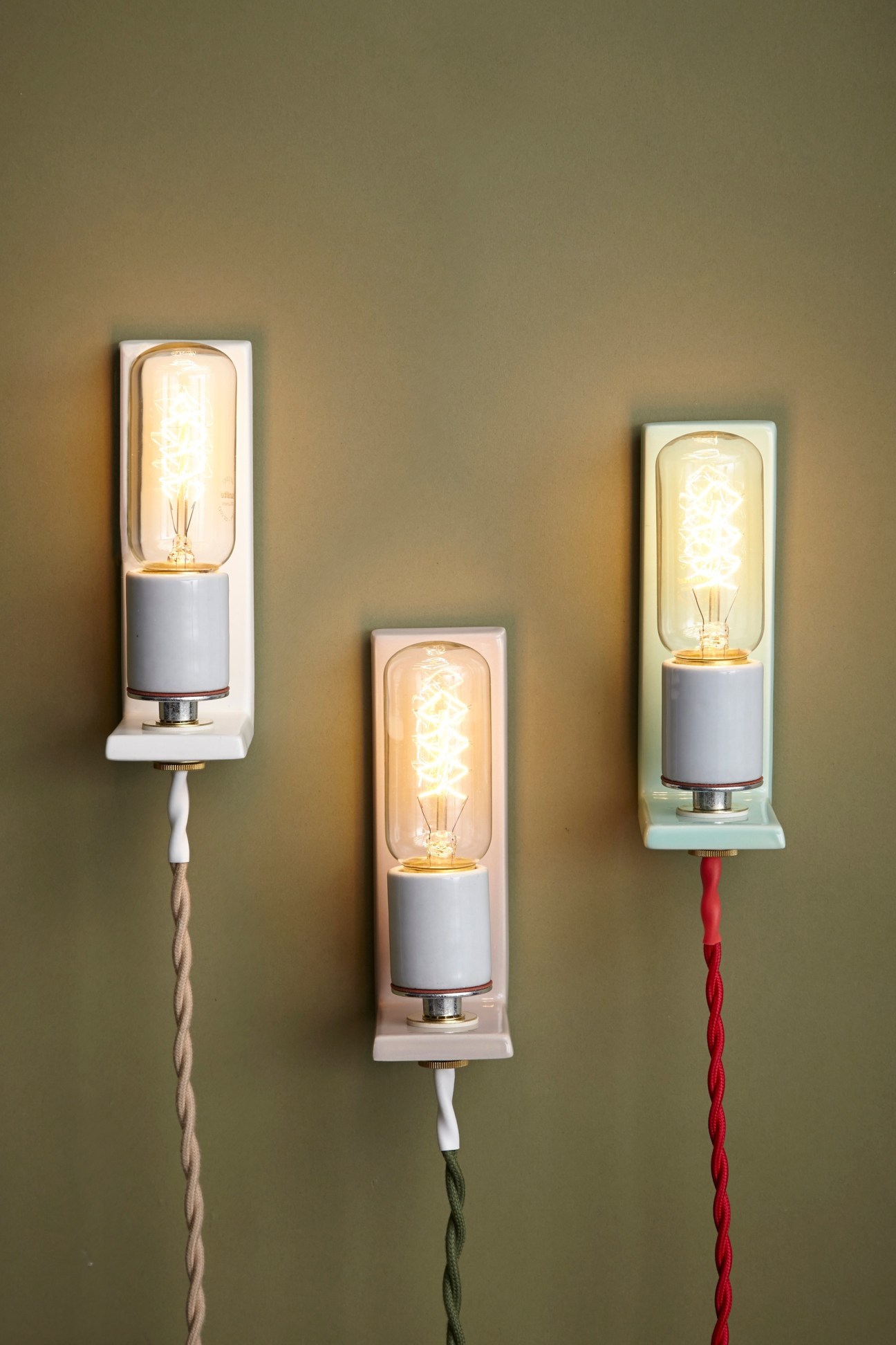 Three L-Lamp wall lights with ceramic bases, porcelain sockets and braided cords in white, sage and mint mounted on a green wall.