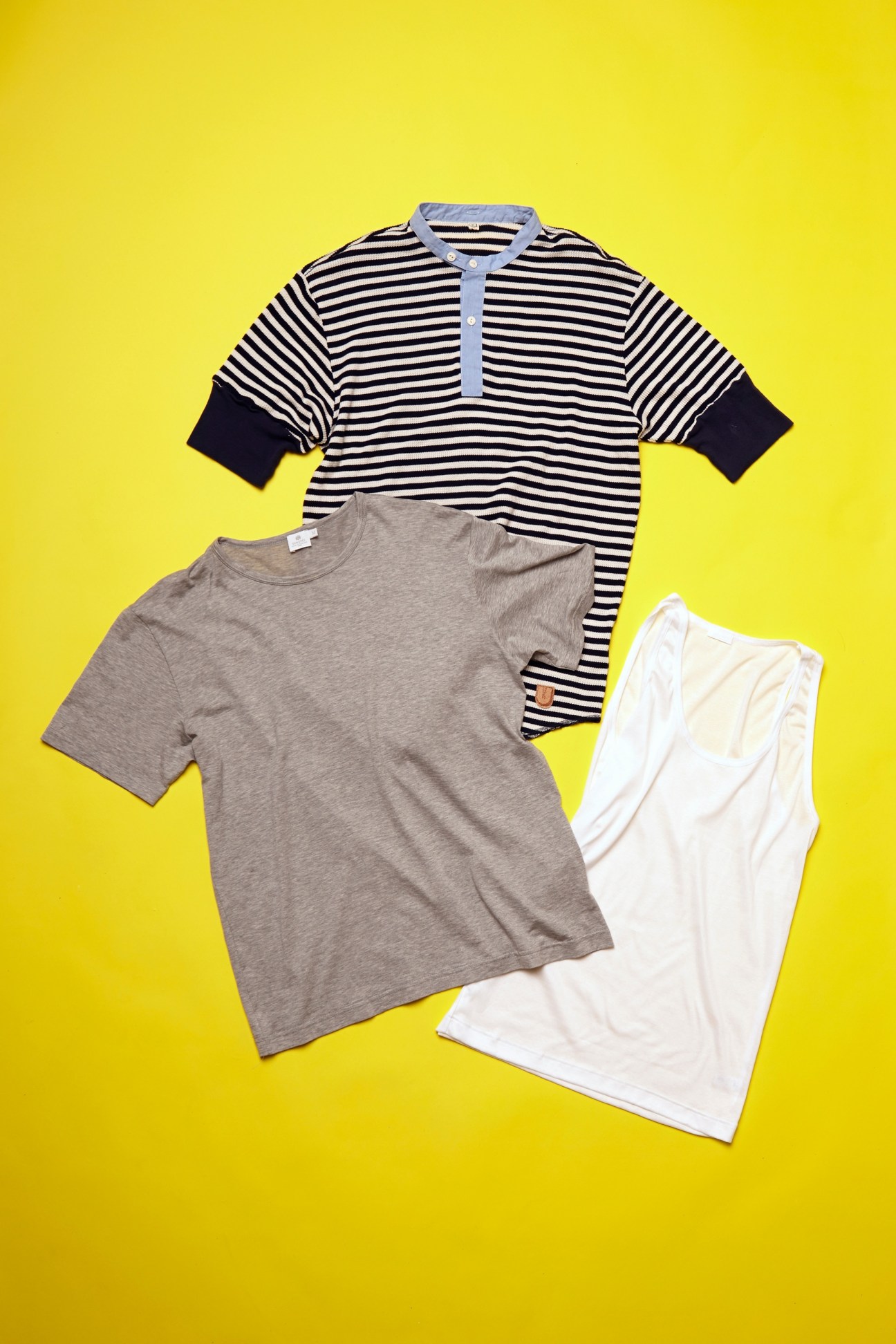 Three t-shirts displayed on yellow background: striped navy and white henley, gray crew neck, and white tank top from UNDER, Sunspel and Zimmerli.
