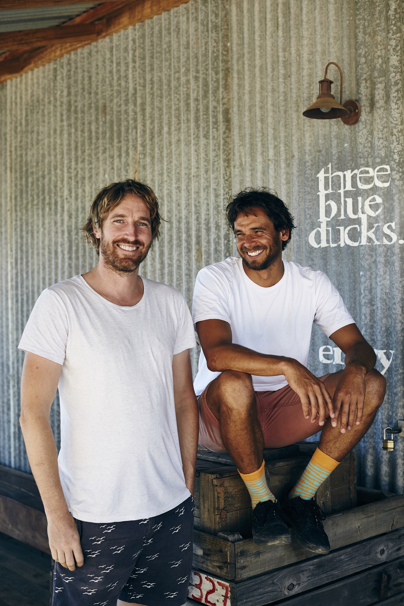 Darren Robertson and Mark LaBrooy, owners of Three Blue Ducks restaurant, smiling in front of corrugated metal wall with rustic lamp.