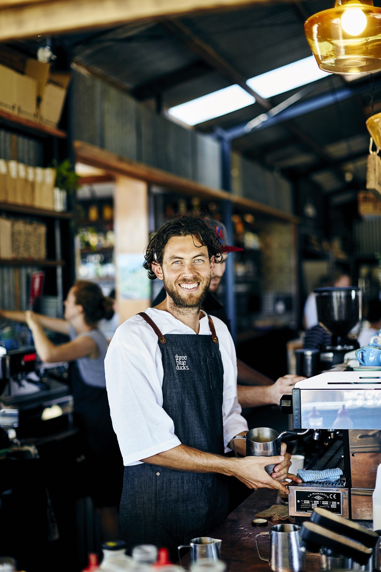 Smiling barista in dark apron at Three Blue Ducks restaurant counter in Byron Bay.