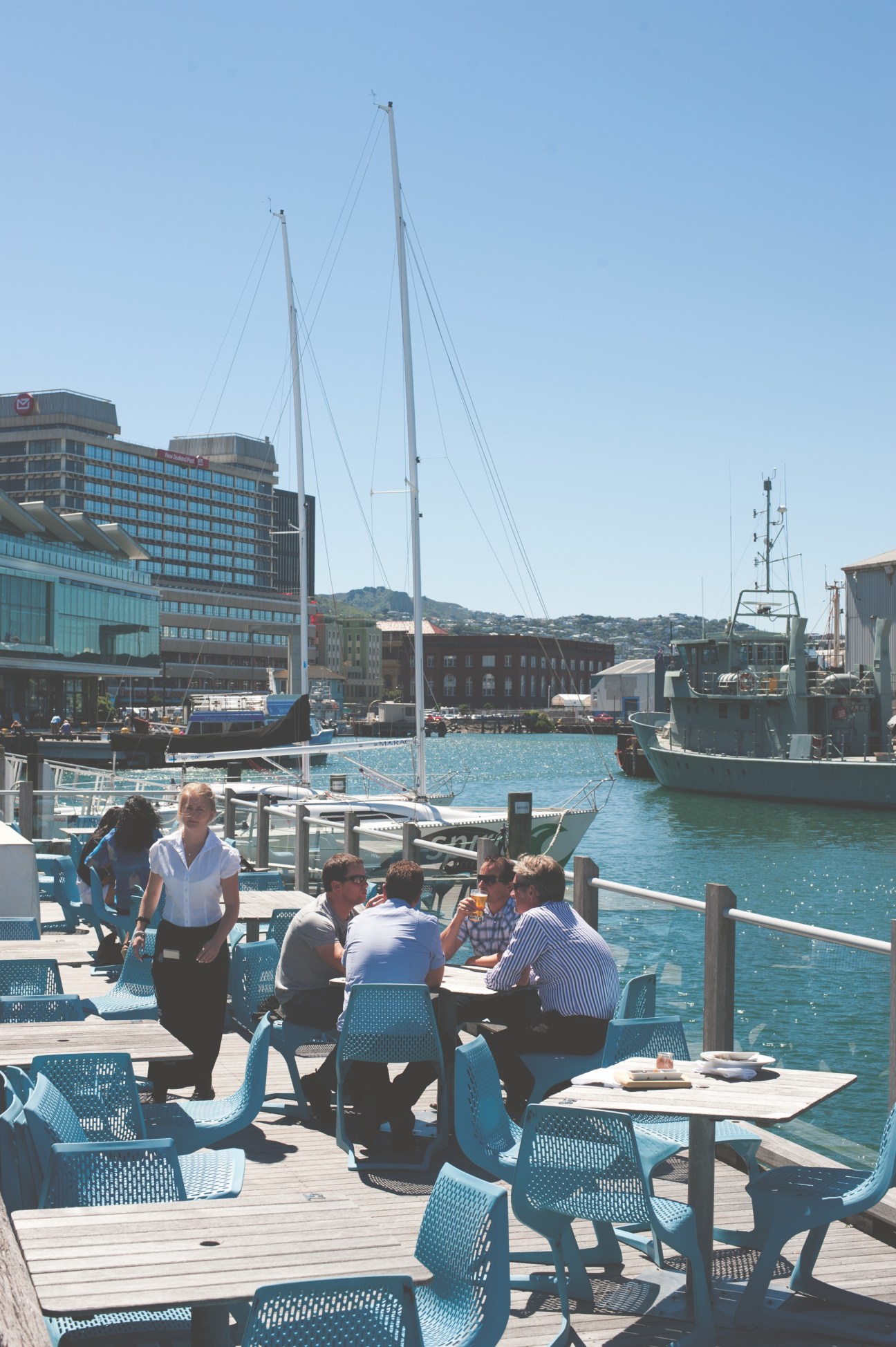 People dining at waterside tables with sailboats and modern buildings in Wellington harbour.