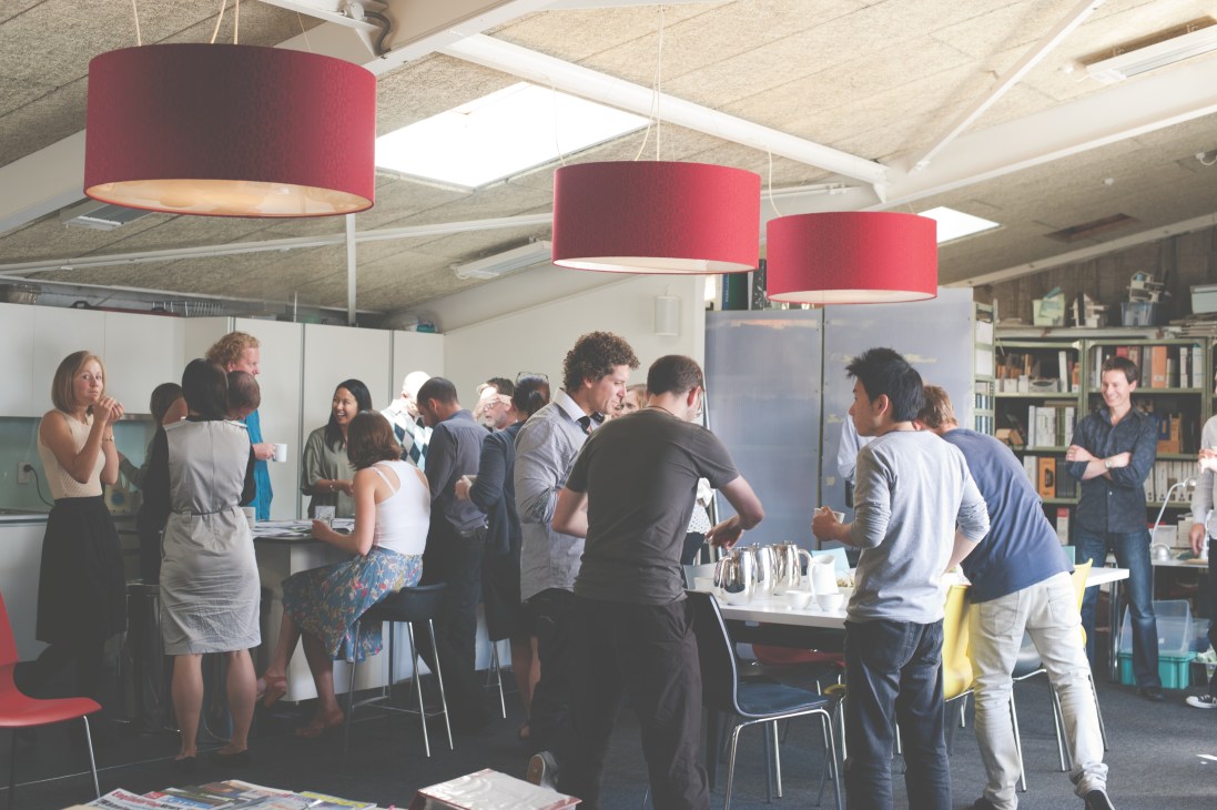 A group of people gather during morning tea at Studio of Pacific Architecture office, standing beneath red pendant lamps.