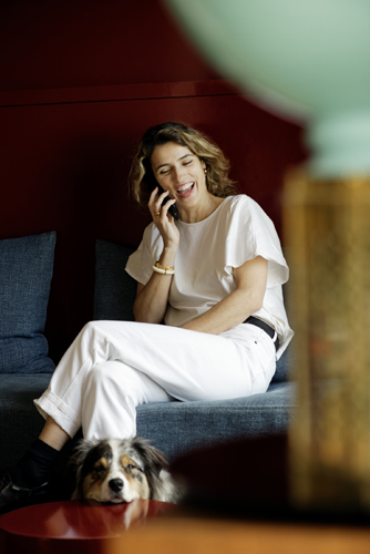 Woman in white clothing sitting on a blue couch while on a phone call, with a dog at her feet.