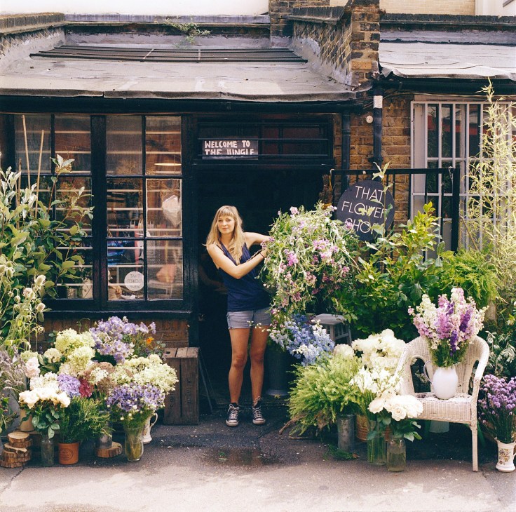 Hattie Fox stands outside That Flower Shop storefront surrounded by colorful seasonal flowers in vases.