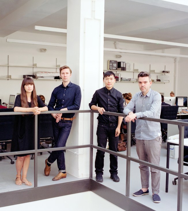 Four design professionals posing at a railing in a modern office space with white walls and shelving.