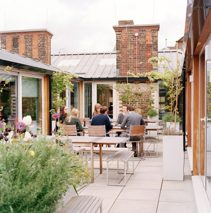 Employees gathered around a wooden table on a rooftop garden terrace with brick chimneys and modern glazed structures.