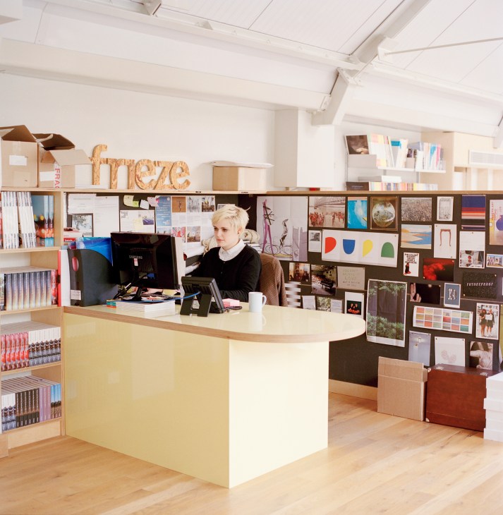 Reception desk at Frieze London office with employee seated at curved yellow and white desk beneath wooden 'frieze' sign.