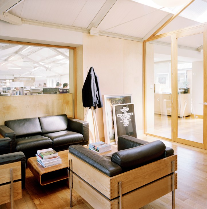 Modern office lounge with black leather seating, wooden furniture, and glass partition walls in a light-filled space.