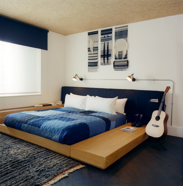 Ace Hotel London bedroom with wooden bed frame, navy headboard, blue quilted bedding, white pillows, wall-mounted lamps, acoustic guitar, and woven wall hangings.