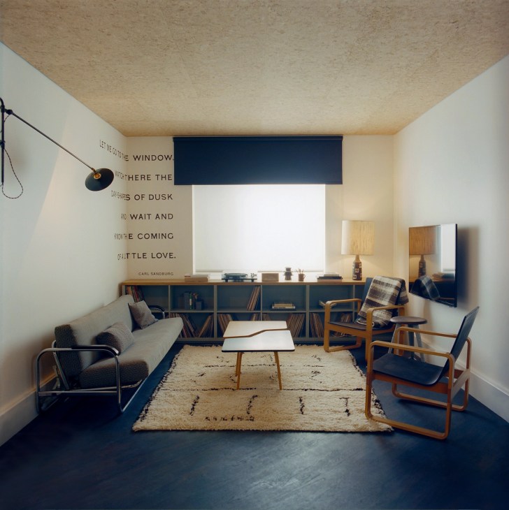 A modern hotel suite sitting room with vintage mid-century furniture, including wooden armchairs, a sofa, and shelving unit beneath a window with inspirational text on the wall.