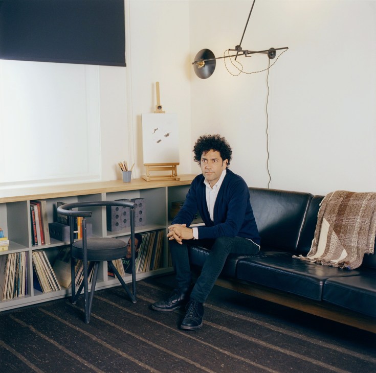 Alex Calderwood sits on a black leather sofa in an Ace Hotel room with minimalist furnishings and industrial lighting.