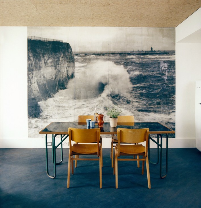 A minimalist interior with wooden chairs at a table beneath a large seascape wall mural featuring white cliffs and crashing waves.