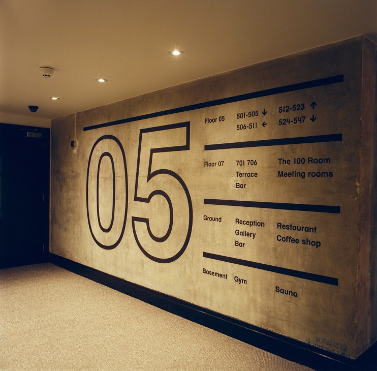 Hotel corridor with large brass-colored wayfinding sign displaying floor numbers and amenity locations in navy text.