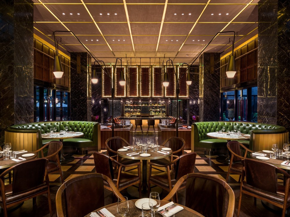 Upscale dining room with green leather banquettes, wooden chairs, geometric ceiling design and illuminated bar.