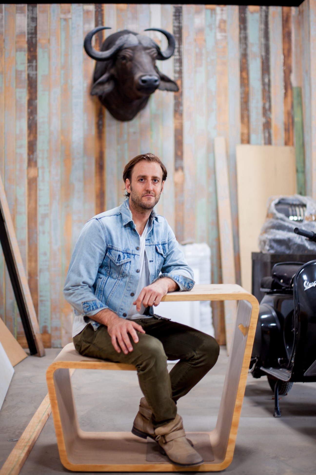 Man in denim shirt sitting on wooden chair in studio with bull sculpture on wall