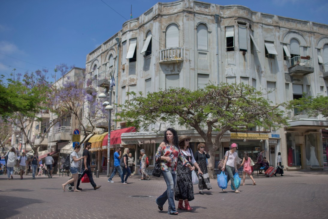 Busy Carmel Market in Tel Aviv with shoppers walking through a pedestrian area lined with historic buildings and purple flowering trees.