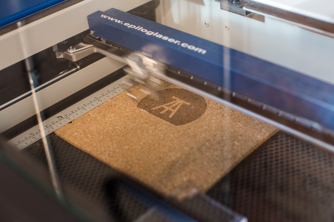 A laser cutter engraves the Atolye logo onto a cork surface inside the machine.