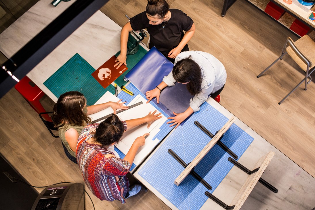 Three people work together on designing desk lamps at a wooden workbench in a bright studio space.