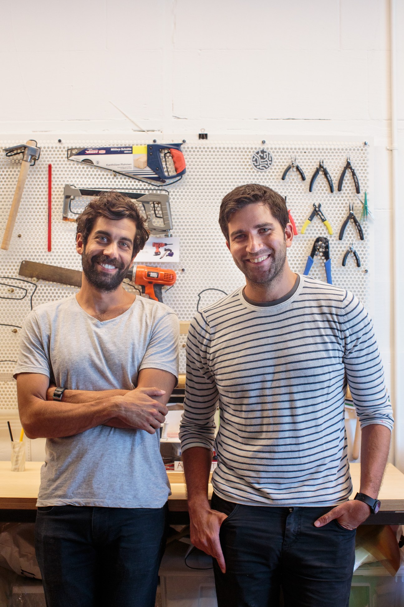 Atolye co-founders Kerem Alper and Engin Ayaz stand smiling in front of a pegboard wall displaying various tools.