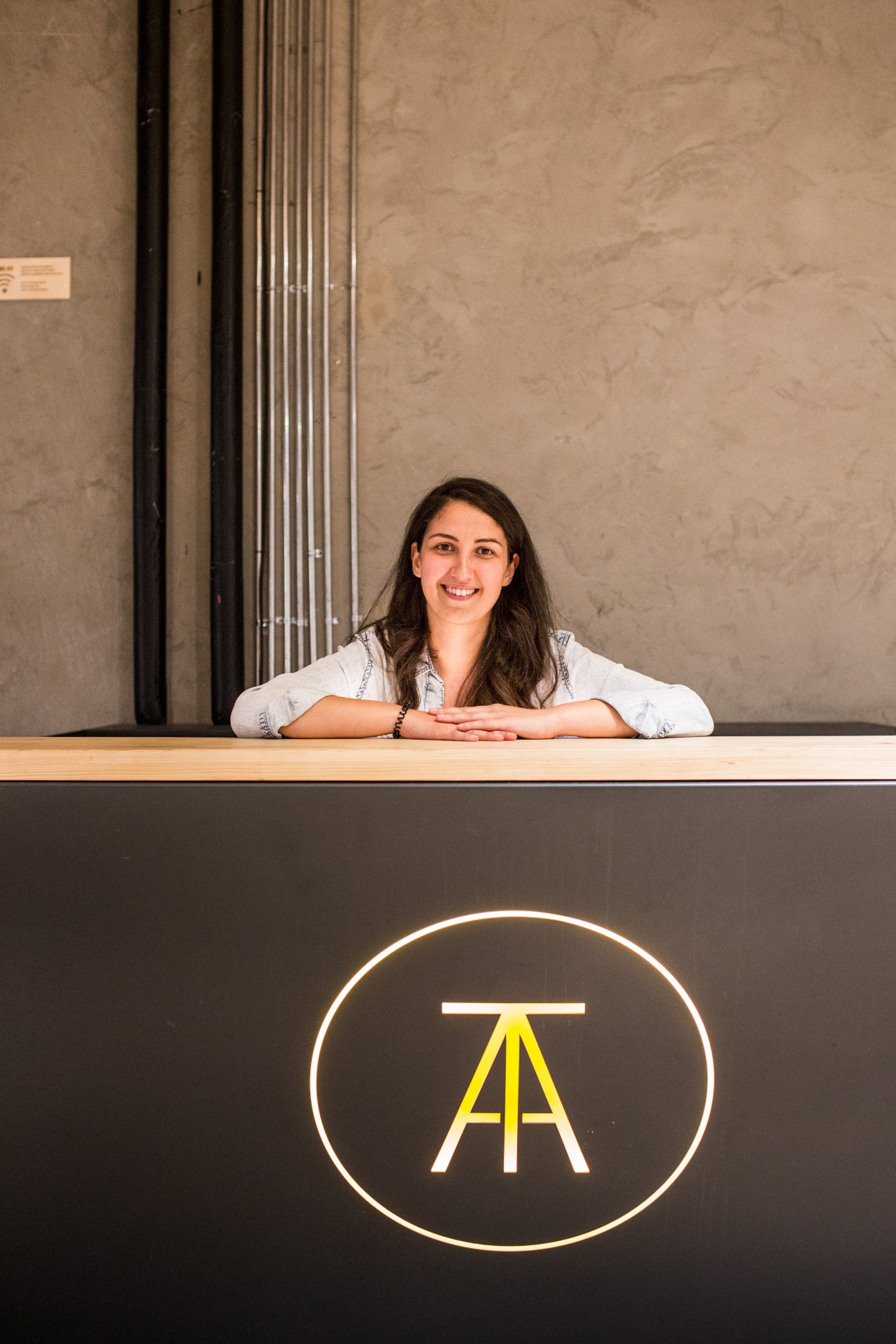 Tugce Karatas, space coordinator at Atolye, stands behind a reception desk with the Atolye logo displayed below.