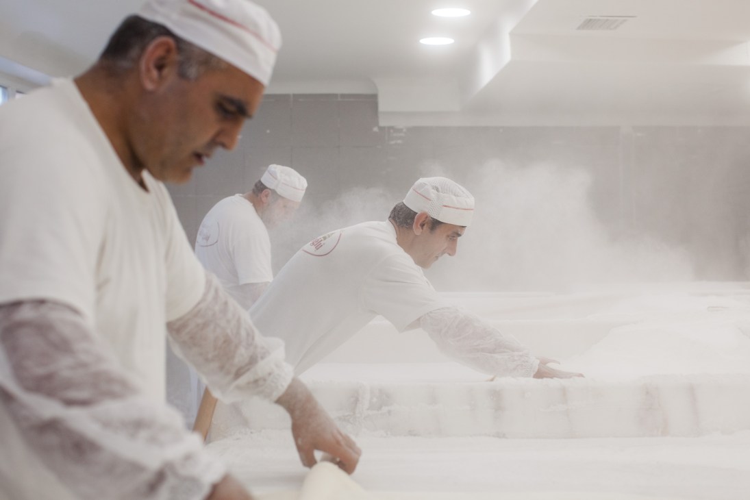 Baklava makers in white uniforms and head coverings work with pastry in a flour-dusted kitchen at Karakoy Gulluoglu.