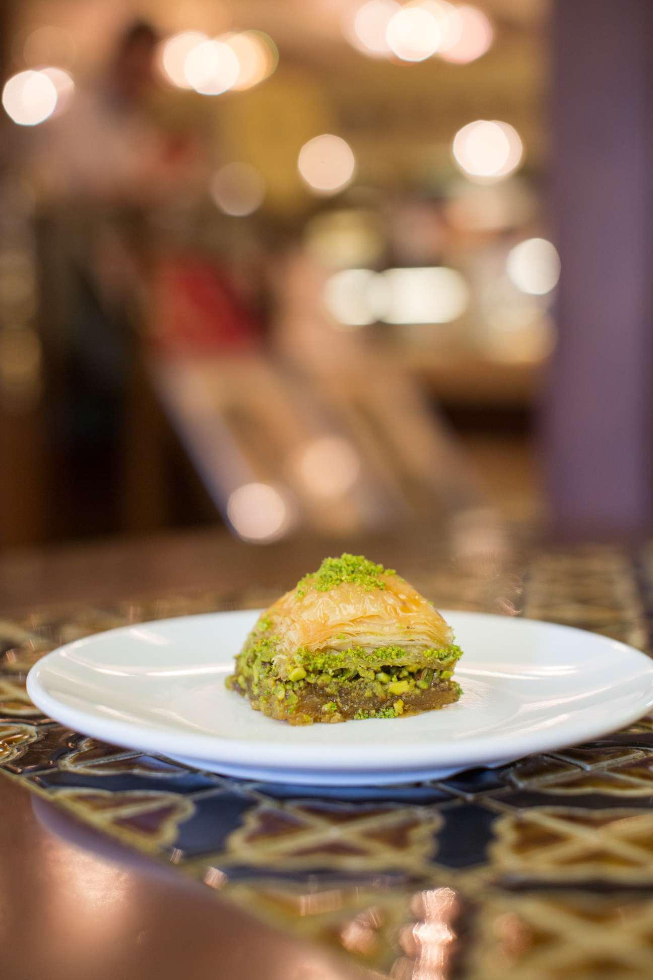 A piece of pistachio baklava on a white plate with blurred background lights and decorative table.