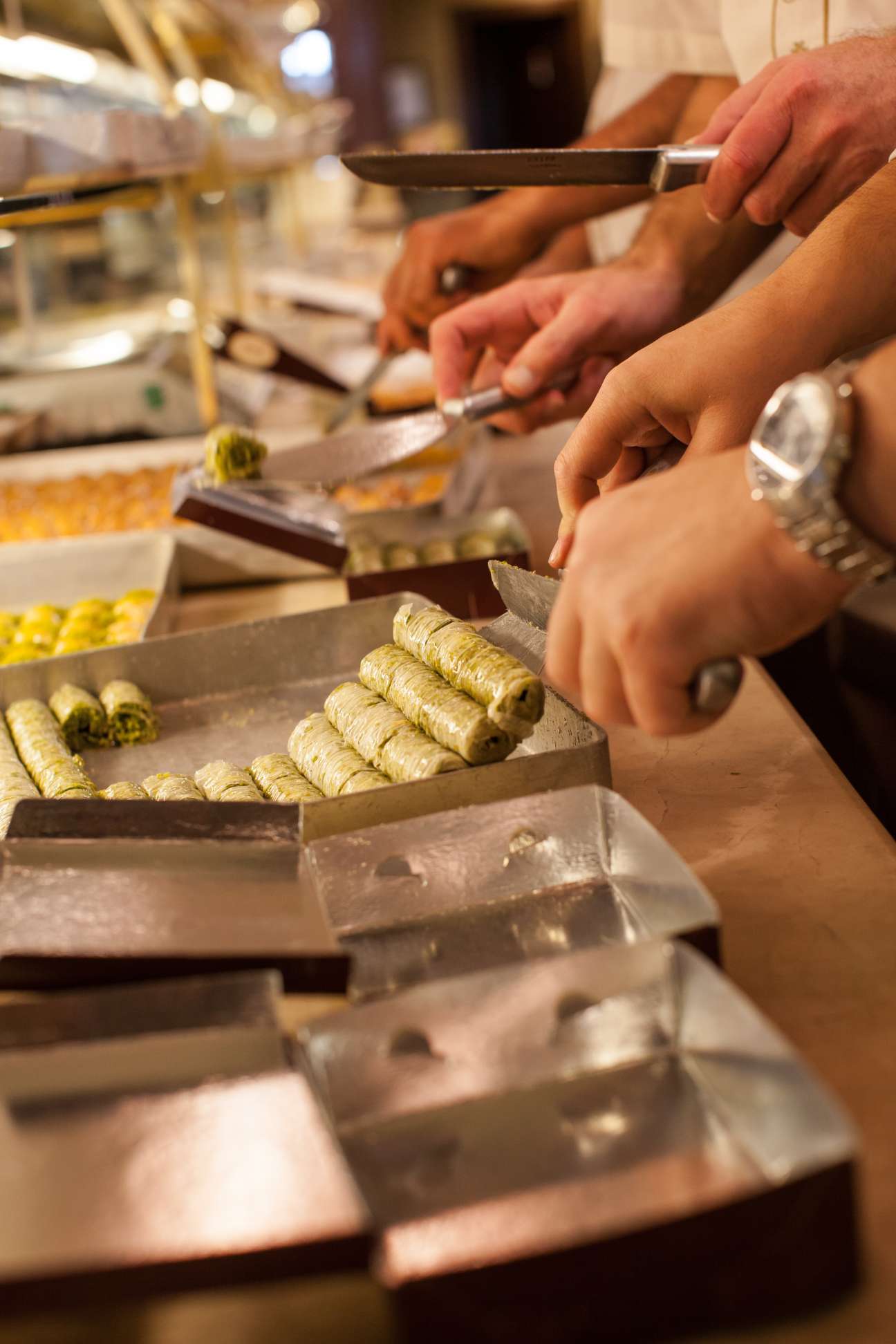 Hands arranging pistachio-filled baklava rolls in metal trays at Karakoy Gulluoglu in Istanbul.