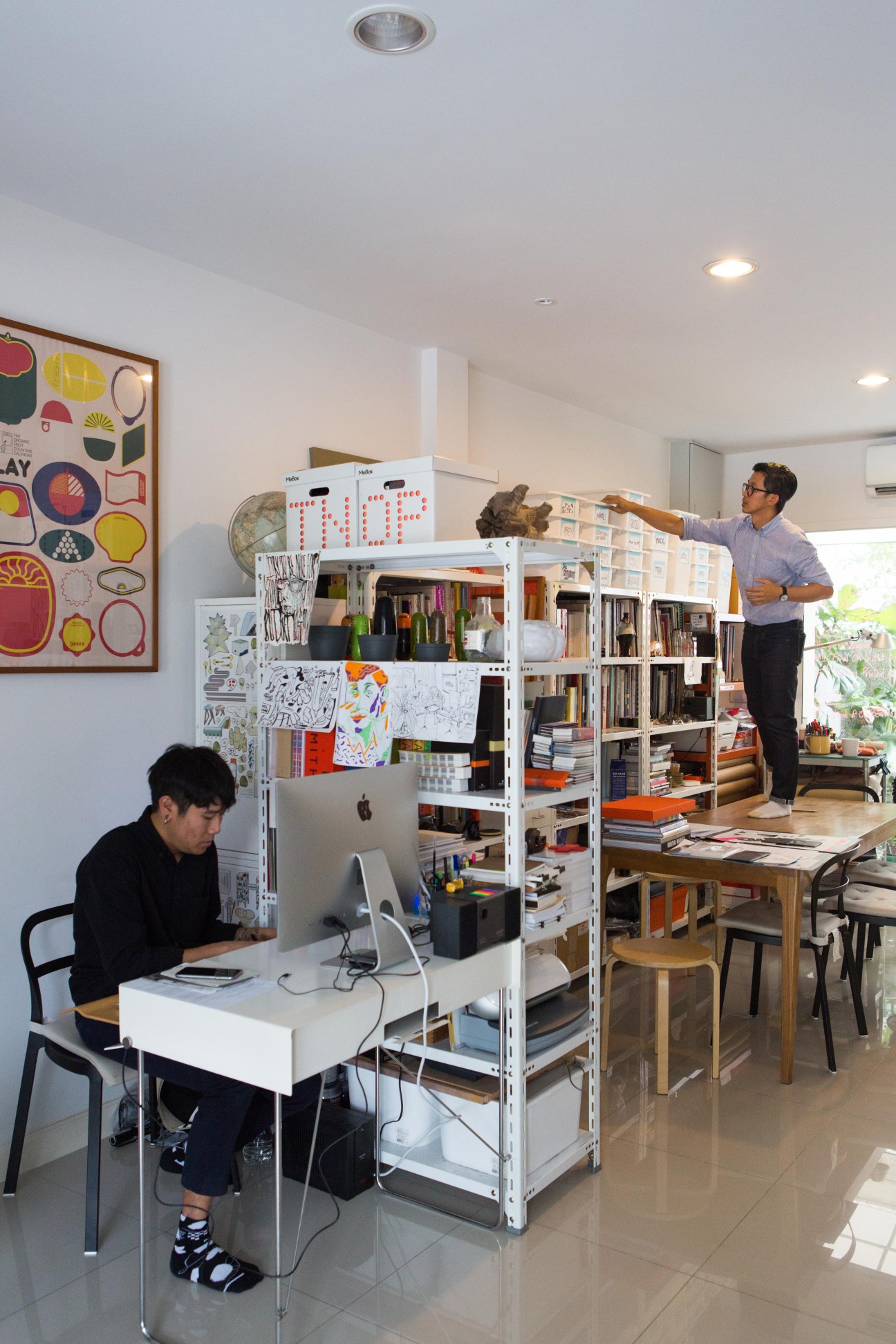 Two men working in a modern design studio with white shelving system containing books and materials