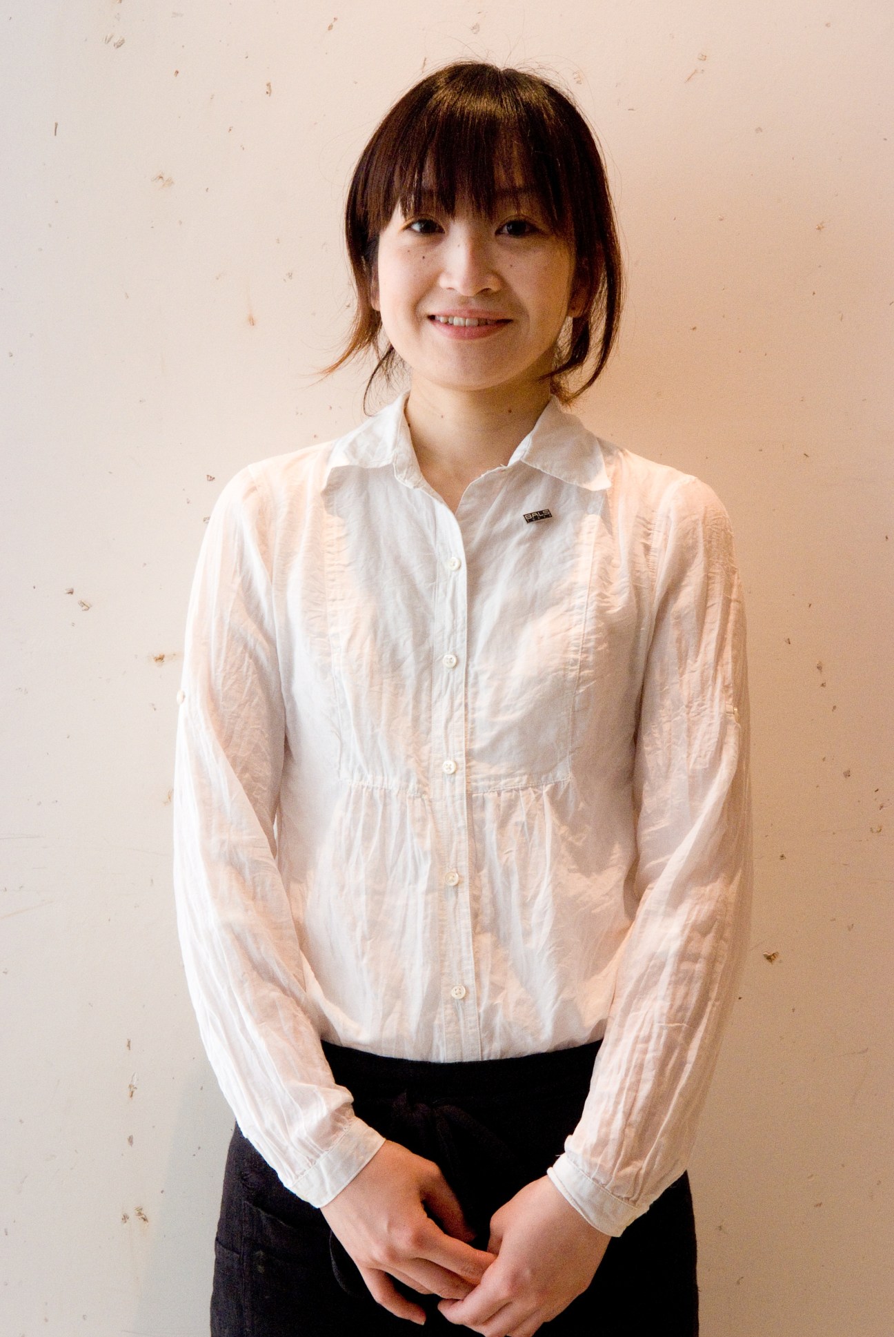 Tomoha Tokushima, a young woman with shoulder-length dark hair wearing a light pink button-up shirt and dark skirt, smiling at the camera.
