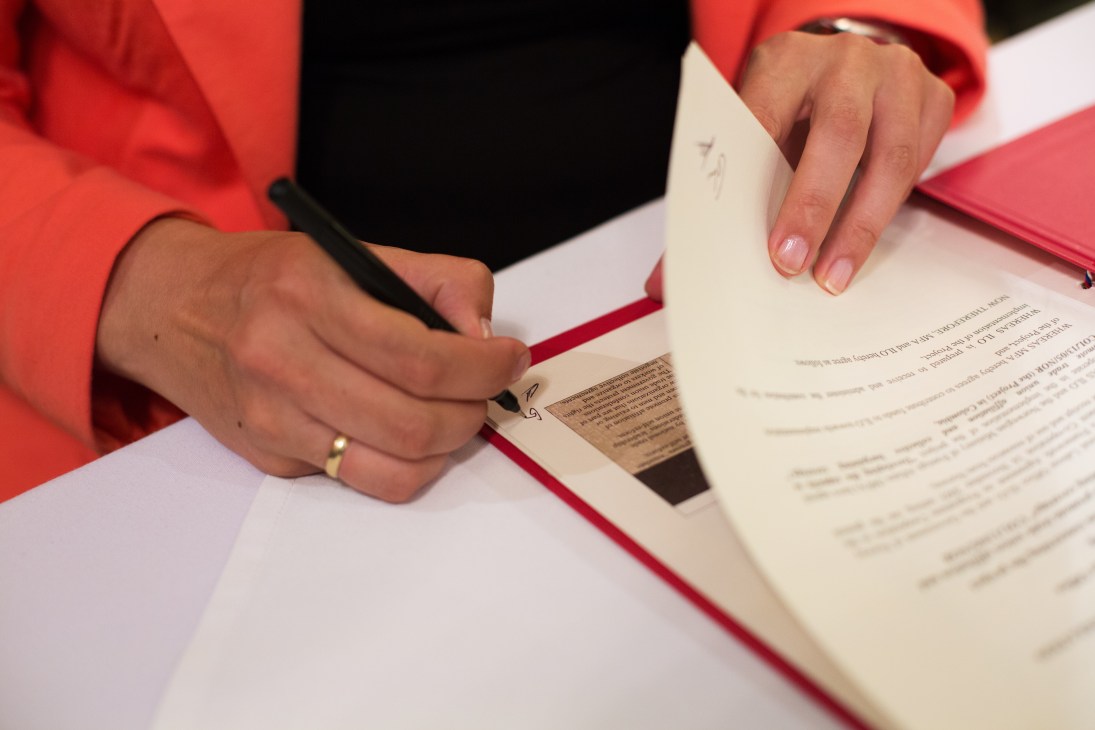 A person in a red jacket signs a document on a table with a red folder.