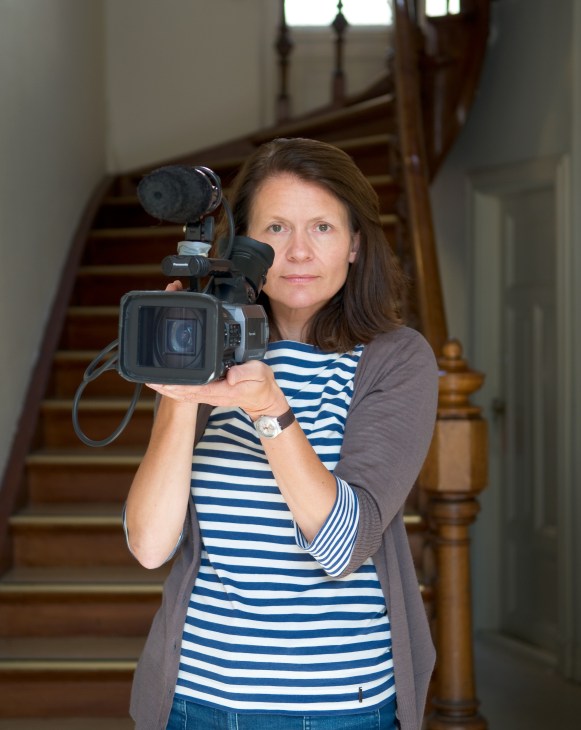Aslaug Holm holding a professional video camera on a wooden staircase.