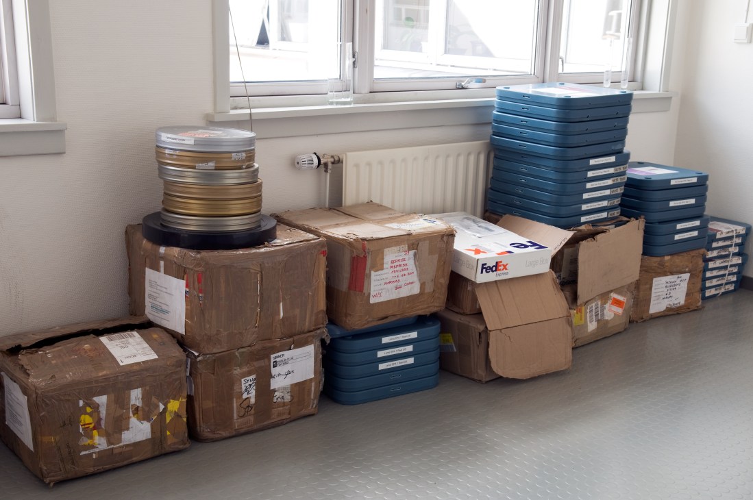 Cardboard boxes and blue film cases containing reels of film stacked in a bright room with large windows.