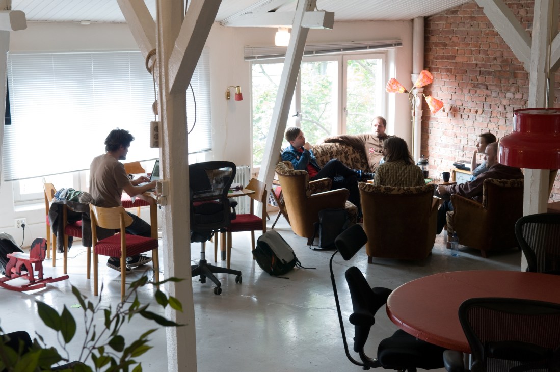 People working in a bright loft-style office space with exposed brick walls, red furniture, and large windows.